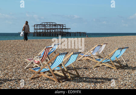 Leere Liegestühle am Strand von Brighton mit Blick auf verlassenen Pier West, Frau zu Fuß am Strand und das blaue Meer in der Ferne Stockfoto