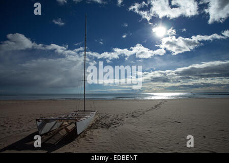 Twin-Rumpf Segelboot an einem Strand mit Sonne in einem wolkenverhangenen Himmel. Fußspuren führen vom Boot auf das Meer. Stockfoto