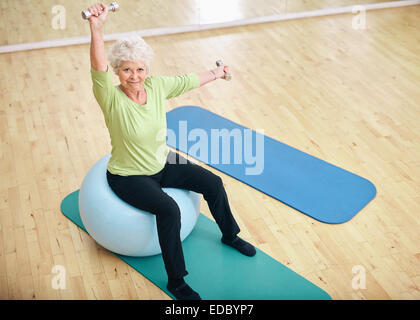 Aktive senior Frau sitzt auf einem Pilates Ball und heben der Hanteln Blick in die Kamera. Alte Frau kaukasischen Training mit wiegen Stockfoto