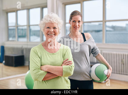 Porträt von glücklich senior Frau mit ihren verschränkten Armen und ihr persönlicher Trainer im Hintergrund Fitnessraum. Fitness im Fitness-Studio. Stockfoto