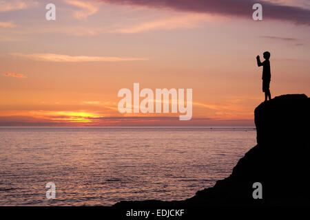 Silhouette der Youngster mit Handy-Kamera auf einem Felsen am Meer bei Sonnenuntergang fotografieren Stockfoto