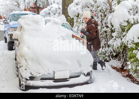 Junger Mann, gekleidet in einen Wintermantel, Hut und Handschuhe reinigt Schnee vom Auto nach Schneesturm mit einem Pinsel. Stockfoto