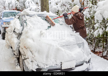 Junger Mann, gekleidet in einen Wintermantel, Hut und Handschuhe reinigt Schnee vom Auto nach Schneesturm mit einem Pinsel. Stockfoto