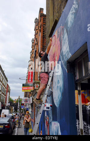 Mann auf einer Leiter malerei Kunst an der Wand und eine Frau mit Kind auf ihren Schultern Spaziergänge durch in Shoreditch, London, England Stockfoto