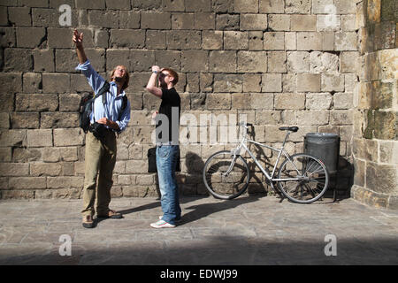 Zwei männliche Touristen vor Steinmauer, nachschlagen, mit dem Fahrrad hinter sich lassen, in mittelalterlichen Marktplatz "Plaça del Rei", Barcelona. Stockfoto