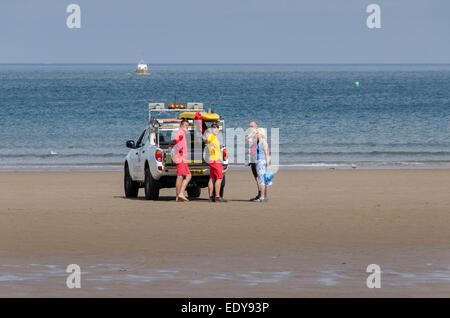 2 männlichen RNLI Lifeguards stehen 4x4 patrol Fahrzeug Sprechen & Beratung zu ein paar Urlauber - Whitby Beach, North Yorkshire, England. Stockfoto