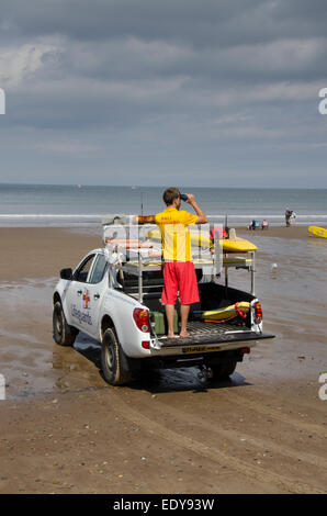 Junge RNLI Rettungsschwimmer Mann auf Pflicht betrachten Menschen mit Fernglas im Meer, steht auf der Rückseite des Pick-up-Truck - Whitby Beach, North Yorkshire, England, UK. Stockfoto