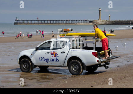 Junger Mann auf Pflicht als rnli Lifeguard ist gerade Menschen in Meer und steht auf der Rückseite des geparkten Pick-up Truck - Whitby Beach, North Yorkshire, England, UK. Stockfoto