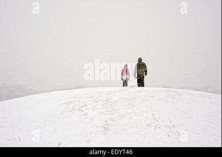 Zwei kleine Figuren in farbenfroher Winterkleidung stehen auf einem riesigen, schneebedeckten Hügel, der von der weiten weißen Landschaft Schottlands im Schatten steht. Stockfoto