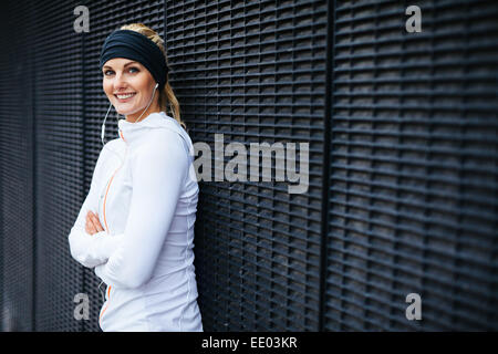 Porträt von schöne junge Frau in Sportkleidung, Blick auf die Kamera zu Lächeln. Sportliche Frau, die an eine Wand gelehnt. Stockfoto