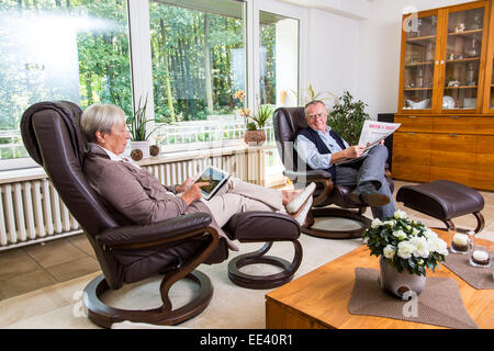 Senior paar, Mitte 70, zu Hause im Wohnzimmer, sie mit einem Tablet PC, iPad, im Internet surft, er liest Zeitung, Stockfoto