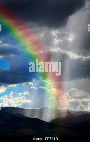 Sonnenlicht Strahlen aus den Wolken fallen auf dunkle Gebirgskette mit Regenbogen Stockfoto