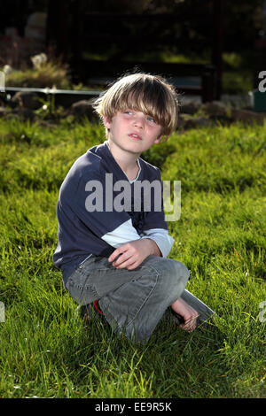 Blonde behaarter Junge sitzt in einem sonnigen Garten und ist in Gedanken versunken Stockfoto