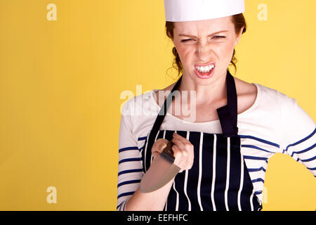 Eine junge und wütend Köchin zeigt ein Kochmesser auf einem gelben Hintergrund. Stockfoto