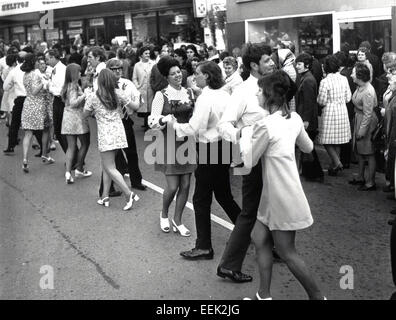 1960er historischen, Shopper, Männer und Frauen zusammen tanzen auf der Straße, mit Mädchen tragen Blumen gemusterte Kleider und kurze Miniröcke, London, England. Possiblly Carnaby Street, dem Zentrum der britische Mode in dieser Ära. Stockfoto