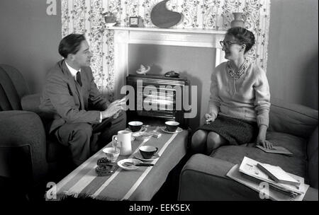 1950er Jahre historische Bild eines jungen Mannes trägt einen Anzug und eine ältere Dame sitzt vor ein elektrischer Kamin im Wohnzimmer mit einer Tasse Tee. Stockfoto