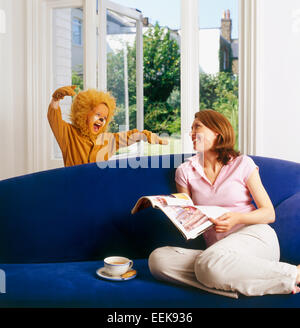 Mama zu Hause entspannen, lesen eine Zeitung, ihr Kind als gekleidet Löwe Überraschungen ihr hinter dem Sofa brüllender erschreckend Stockfoto