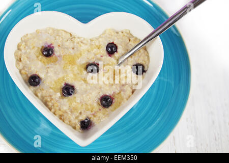 Heidelbeer-Porridge oder Haferflocken mit Honig in eine herzförmige Schüssel mit einem Löffel. Ruht auf der blauen Platte auf einem weißen Hintergrund rustikal. Stockfoto