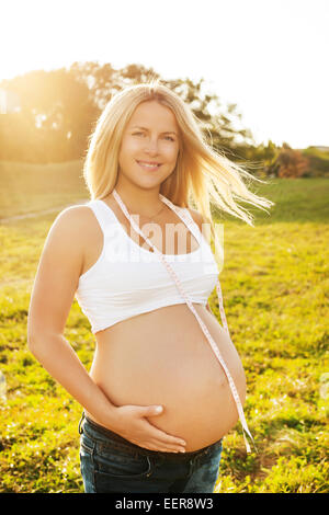 Schöne blonde schwangere Frau im Sommerpark in der Sonne stehen, halten Maßband. Natürlichen Hintergrund Stockfoto