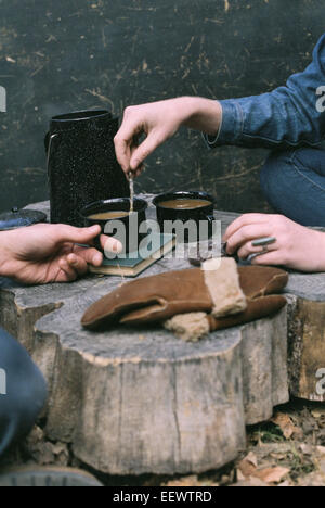 Paar beim Kaffee in einem Wald, zwei Tassen Kaffee, eine Kaffeemaschine und ein paar Handschuhe auf einem Baumstamm. Stockfoto