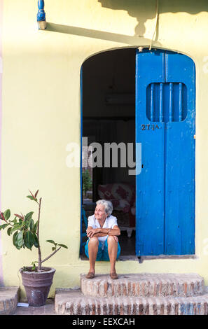 Lokale ältere Frau friedlich sitzen entspannt auf der Haustür in eine Tür von einem gelben Haus mit blauen Türen in Camagüey, der drittgrößten Stadt Kubas Stockfoto