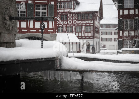 Brunnen mit Schnee und Eiszapfen mit typisch Schweizer Bar befindet sich im Hintergrund, Bülach, Schweiz. Stockfoto