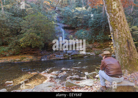 Ein Mann trägt eine Lederjacke und Aussie Hut sitzt auf einem Felsen mit Blick auf einen malerischen Wasserfall und Stream im Herbst Stockfoto