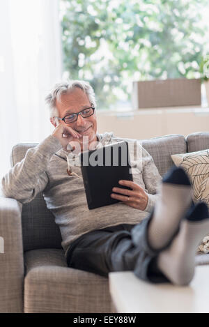 Ältere Mann sitzt auf der Couch im Wohnzimmer, TabletPC verwenden und lächelnd Stockfoto