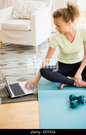 Junge Frau sitzt auf der Yogamatte und mit Laptop im Wohnzimmer Stockfoto