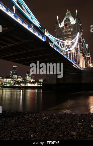 Blick auf die Tower Bridge von unten bei Nacht. Die ferne Skyline Londons über die Themse, umrahmt von der Unterseite der Brücke. London, Großbritannien Stockfoto