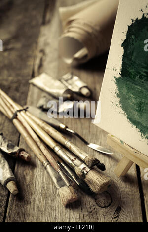 Künstlerische Pinsel, Tuben Ölfarbe, Spachtel und Staffelei mit Ölgemälde auf alten Schreibtisch aus Holz. Vintage stilisierte Foto. Stockfoto