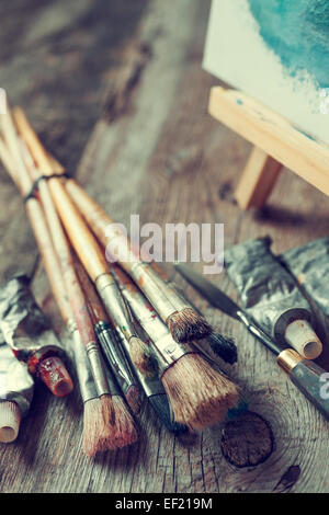 Künstlerische Pinsel, Tuben Ölfarbe, Spachtel und Staffelei mit Ölgemälde auf alten Schreibtisch aus Holz. Stockfoto
