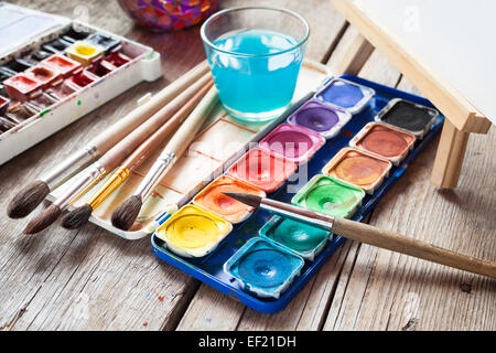 Box mit Aquarellfarben, Pinsel, Glas Wasser und Staffelei mit Leinwand oder Papier auf alten Holztisch. Stockfoto