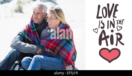 Zusammengesetztes Bild des glücklichen Paares eingehüllt in Decke am Strand sitzen Stockfoto