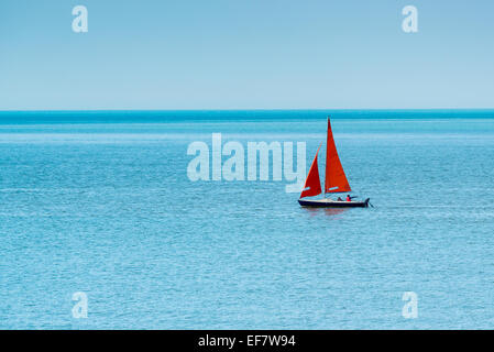 Einzelne Jolle mit rot-Orange-Segeln auf Ruhe, Azure, Meer unter strahlend blauem Himmel. Stockfoto