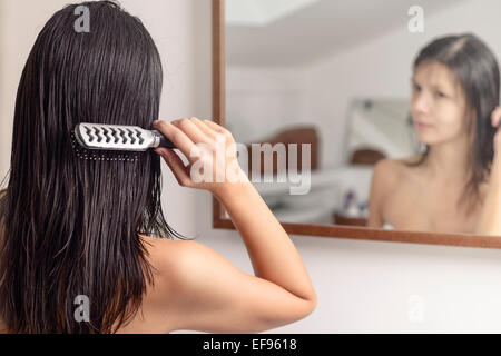 Junge Frau auf der ständigen Suche auf ihr Spiegelbild im Badezimmerspiegel beim Zähneputzen mit einer Haarbürste ihr nassen Haares nach dem Waschen Stockfoto