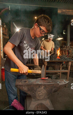 Flammen steigen als moderne Amateur Mutter und Sohn Schmiede eine traditionelle Kohle-Schmiede schüren, während der Arbeit an glühendes Eisen auf einem Amboss an eine Schmiede 1890-Ära an das Orange County Heritage Museum in Santa Ana, CA. Hinweis Schutzbrille und Hammer. Stockfoto