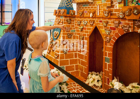 Ein Kind Leben Specialist bei CHOC Kinderkrankenhaus in Orange, CA, weist darauf hin ein Bär-förmigen Lebkuchen auf ein riesiges Lebkuchenhaus in der Klinik-Lobby, ein junges Mädchen-Chemotherapie-Krebs-Patienten. Beachten Sie die Glatze. Kind Leben Spezialisten unterstützen Patienten und Familien im Krankenhausumfeld. Stockfoto