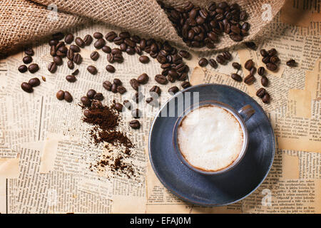 Blaue Keramik Tasse Cappuccino mit gerösteten Kaffeebohnen über alte Zeitung. Ansicht von oben. Stockfoto