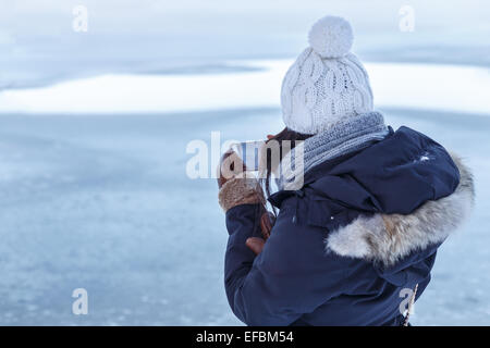 Junge asiatische Frau in Winterkleidung von hinten zu sehen, wie sie ein Foto auf ihrem Smartphone vor einem zugefrorenen See blickt. Stockfoto