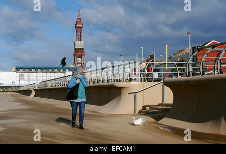 Eine Frau, die gegen die kalte Spaziergänge entlang Blackpool Promenade gewickelt, wie der Sand geblasen wird oben durch den Wind im Winter Wetter UK Stockfoto