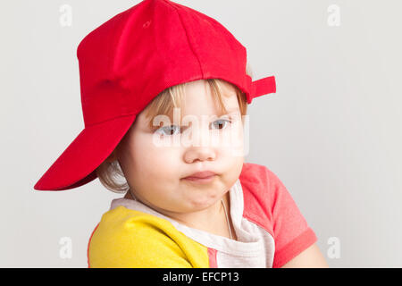 Studioportrait lustig verwirrt Baby girl in rote Baseballmütze über graue Wand Hintergrund Stockfoto