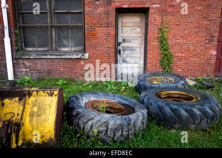 Alte, verlassene Ausrüstung und großen Reifen sitzen unter Rasen und Unkraut vor einem roten Backstein-Gebäude. Stockfoto