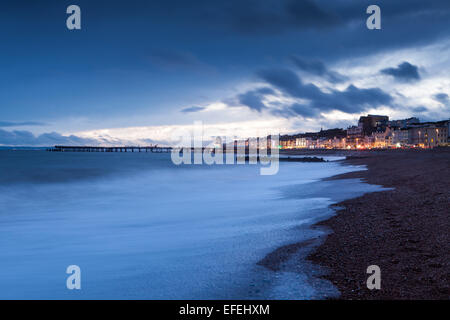 Hastings Strandpromenade und Pier. Wellen auf den Strand bei Sonnenuntergang. während die Lichter der Stadt funkeln im Hintergrund. Stockfoto