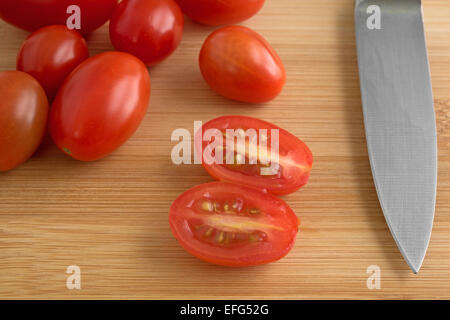 Ein einzelnes schneiden Traube Tomaten neben einem Messer mit mehr Tomaten im Hintergrund auf einem Holz Schneidebrett Stockfoto