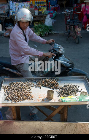 Ein Mann auf einem Motorrad blickt auf ein Tablett mit Schnecken als Straße Nahrung auf einer Stadtstraße in Kampong Cham, Kambodscha erhältlich. Stockfoto