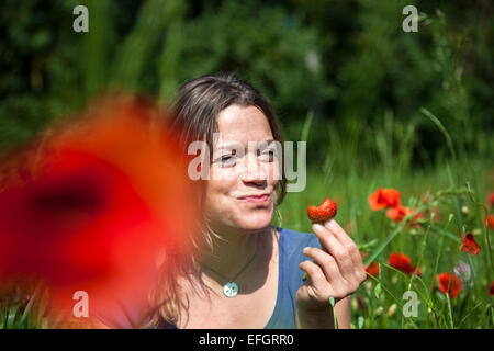 Junge Frau sitzt in einem blühenden Mohn-Feld eine Erdbeere essen Stockfoto