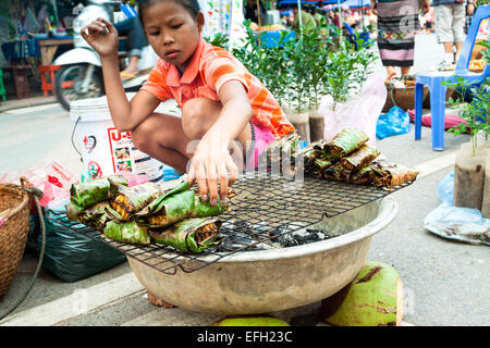 Ein kleines Mädchen, Verkauf von gegrilltem Waben, eingewickelt in Bananenblätter am Morgenmarkt in Luang Prabang, Laos. Stockfoto