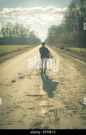Eine Dame mit Koffer warten allein in der Mitte einer Landstraße, die durch Felder und Wälder. Schuss bei Gegenlicht. Stockfoto