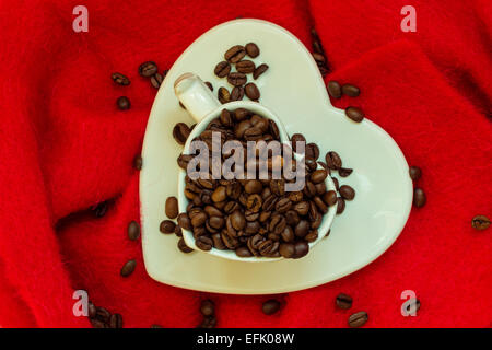 Kaffee Klatsch Java-Konzept. Herzförmige weiße Tasse gefüllt mit gerösteten Kaffeebohnen auf rotem Tuch Hintergrund Stockfoto